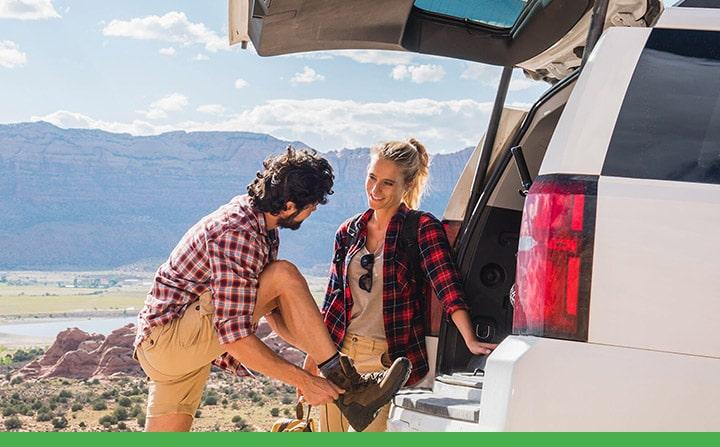 Your Car a couple lacing up hiking boots in the mountains behind the open trunk of an SUV